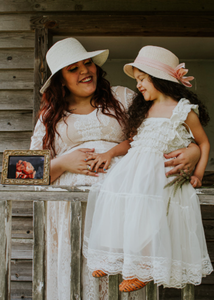 Mother 30 with daughter 8. Both in white frilly dresses. both wear white floppy hats. daughter has hand on mothers pregnant belly  