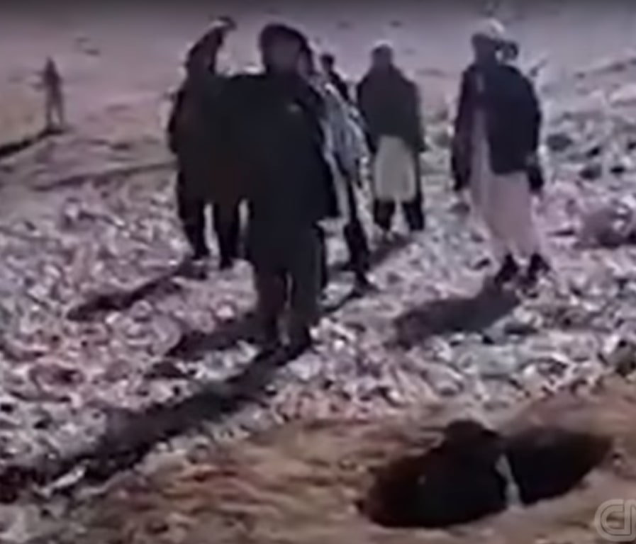 A person is in a hole buried to her shoulders. desolate location. 7 men wearing tribal desert clothing. one mas is in the motion of throwing a stone